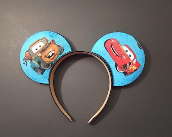 May include: A pair of mouse ears featuring the characters Mater and Lightning McQueen from the movie Cars. The ears are round with a light blue background and black beaded trim. The headband is tan.