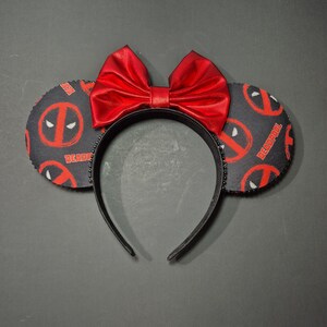 May include: A headband featuring two circular ears covered in a black fabric with a repeating red Deadpool logo pattern and the text Deadpool. A large, shiny red bow is centered between the ears, and the headband band is black with small studs.
