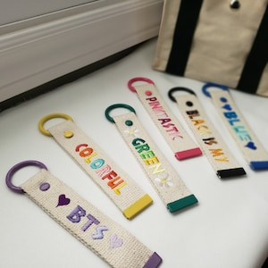 Puede incluir: Seis llaveros coloridos con texto bordado sobre un fondo de lona blanco. Los llaveros presentan las palabras "Pinktastic", "Green", "Colorful", "BTS", "Black is My" y "Blue".