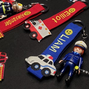 May include: Three personalised keychains with embroidered names, a red fire engine, a white ambulance, and a white police car. Each keychain features a small plastic figurine of a firefighter, a paramedic, and a police officer.