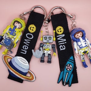 Peut inclure: Deux porte-clés personnalisés avec des lanières noires, chacun avec un nom en blanc. L'un a un smiley jaune et le nom "Owen", avec une figurine d'astronaute et un écusson de planète. L'autre a un smiley et le nom "Mia", avec un écusson de fusée et une figurine d'astronaute.
