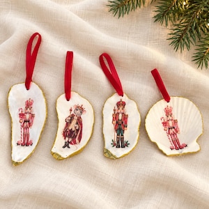 May include: Four white seashell ornaments with gold-painted edges and red ribbon loops. Each shell features a detailed illustration of a nutcracker or a mouse dressed in royal attire, holding a candy cane or a scepter, for holiday decor.