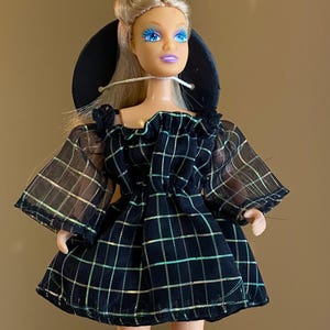Puede incluir: Una muñeca con cabello rubio y ojos azules que lleva un vestido a cuadros negro con mangas transparentes. La muñeca tiene un sombrero negro y posa sobre un fondo neutro. El vestido presenta un patrón de cuadrícula con acentos iridiscentes.