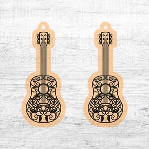 May include: Two wooden guitar ornaments with intricate floral designs and black outlines. The guitars are on a white wood background.