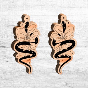 May include: A pair of wooden earrings with a black snake and flower design. The snake is curled around the flower and has a star-like pattern.