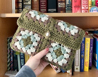 May include: A handmade crochet clutch in shades of olive green, white, light blue, and brown. The granny square pattern is accented with a decorative clasp. The folded design reveals the interior. A stylish accessory for carrying essentials.