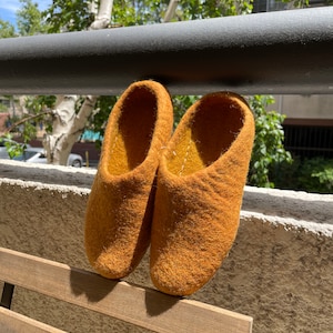 May include: A pair of mustard yellow wool slippers hanging on a wooden railing. The slippers are made of a soft, fuzzy material and have a rounded toe.
