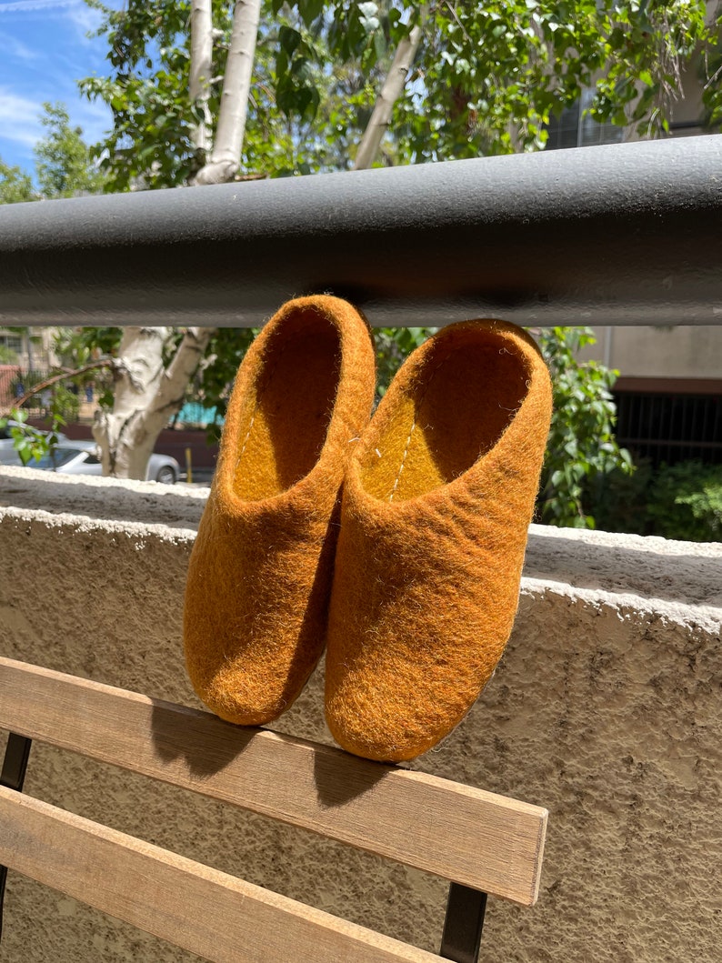 May include: A pair of mustard yellow wool slippers hanging on a wooden railing. The slippers are made of a soft, fuzzy material and have a rounded toe.