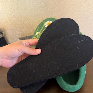 May include: A pair of black felt insoles held in front of a green slipper with a daisy design. The insoles are oval-shaped and appear to be made of a soft, textured material. The slipper is green with a white daisy and yellow center.