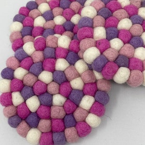 May include: Three round felt coasters in shades of pink, purple, and white. Each coaster is made of small, densely packed felt balls.