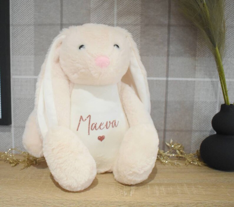 May include: A soft, plush pink bunny with long white ears. The bunny has a white patch on its chest with the name "Maeva" embroidered in pink.
