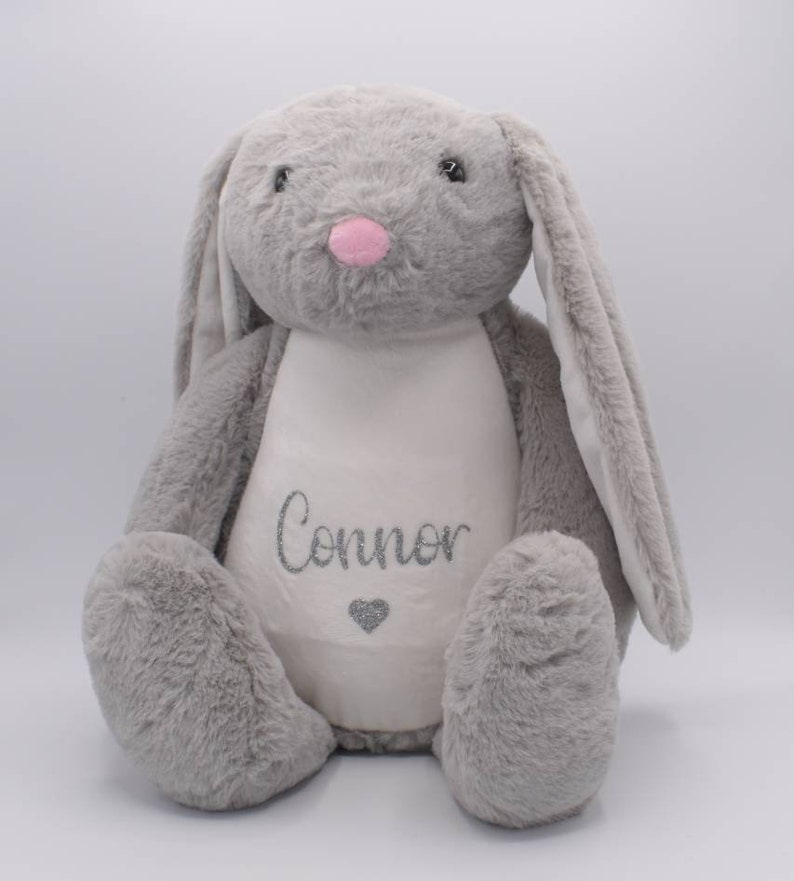 May include: A gray plush bunny with long ears and a white patch on its chest. The patch has the name "Connor" embroidered in silver glitter and a small silver glitter heart.