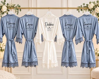May include: Five satin robes hanging on a gold rack. Four are dusty blue and one is white. Each robe features lace trim on the sleeves and hem, with custom black text on the back including names, roles like Bridesmaid or Bride, and the date 14.07.22.