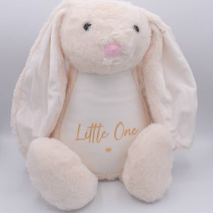 May include: A plush cream-colored bunny with long floppy ears. The bunny is sitting with its paws in front of it. The bunny has a white patch on its chest with the words "Little One" written in gold lettering.