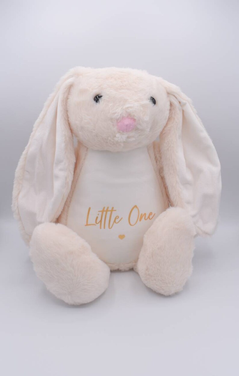 May include: A plush cream-colored bunny with long floppy ears. The bunny is sitting with its paws in front of it. The bunny has a white patch on its chest with the words "Little One" written in gold lettering.