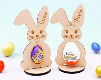 May include: Two wooden bunny-shaped Easter egg holders, one with the name "Luca" and the other with the name "Olivia". Each bunny is holding a chocolate egg, one is a Cadbury Creme Egg and the other is a Kinder Surprise egg.