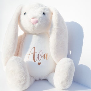 May include: White plush bunny with long ears, sitting with its paws in front of it. The bunny has a pink nose and blue eyes. The bunny has the name "Ava" embroidered on its chest in gold lettering with a small gold heart below the name.