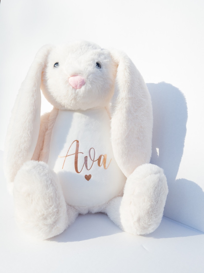 May include: White plush bunny with long ears, sitting with its paws in front of it. The bunny has a pink nose and blue eyes. The bunny has the name "Ava" embroidered on its chest in gold lettering with a small gold heart below the name.