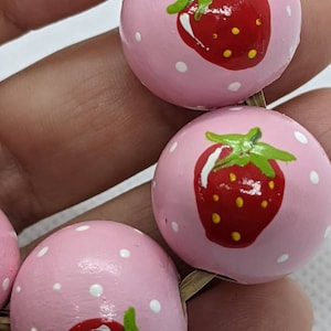 May include: A close-up of a pink wooden bead with white polka dots and a painted red strawberry. The bead is part of a string of beads with a similar design.