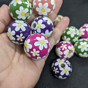 May include: A collection of wooden beads painted with white daisies. The beads are in various colours, including green, pink, purple, and magenta.