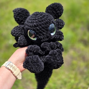 Può includere: Un peluche a forma di drago nero fatto a mano all'uncinetto, con grandi occhi riflettenti. Il drago ha ali dettagliate e una coda con la punta rossa. Il peluche è tenuto su uno sfondo verde sfocato, mostrando la morbida consistenza.