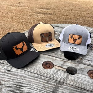 May include: Three trucker hats with a wooden patch featuring a deer head. The hats are black, tan, and gray with mesh backs and the word "KAMEL" on the brim.