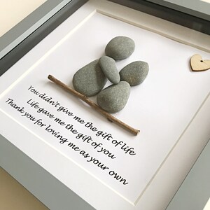 May include: Framed pebble art with a grey frame. The artwork depicts a person made of grey pebbles, a wooden stick, and a small wooden heart. The white background has a printed message in black text.