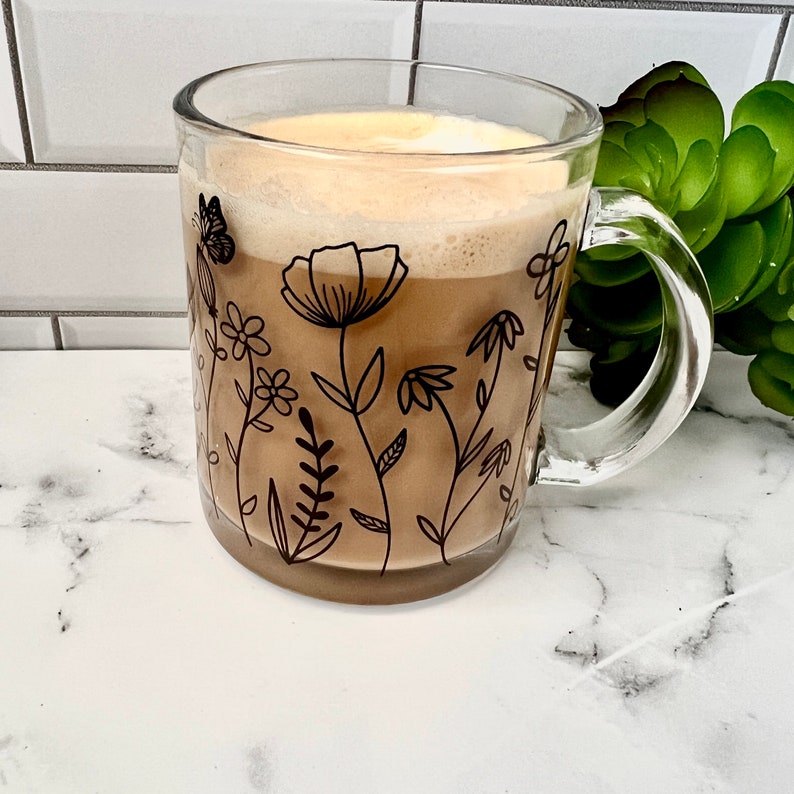 May include: A clear glass mug with a black floral design and a brown coffee drink inside. The mug has a handle and is sitting on a white countertop.