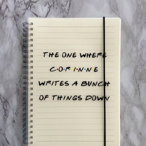 May include: Cream-coloured spiral-bound notebook with lined pages and a black elastic band. The cover displays the text "THE ONE WHERE CORINNE WRITES A BUNCH OF THINGS DOWN" in black, with the name in colourful letters.