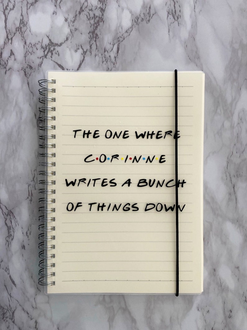 May include: Cream-coloured spiral-bound notebook with lined pages and a black elastic band. The cover displays the text "THE ONE WHERE CORINNE WRITES A BUNCH OF THINGS DOWN" in black, with the name in colourful letters.