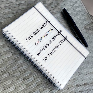 May include: A white, spiral-bound notebook with lined pages and a black elastic closure. The notebook has the text "THE ONE WHERE CORINNE WRITES A BUNCH OF THINGS DOWN" in black and colourful lettering. A black pen is placed on the right.