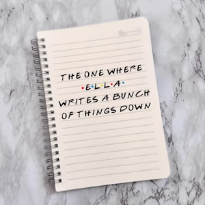 May include: A cream-coloured, spiral-bound notebook with lined pages. The text on the page reads "THE ONE WHERE E.L.L.A. WRITES A BUNCH OF THINGS DOWN" in black ink. The notebook is on a marble-effect surface.