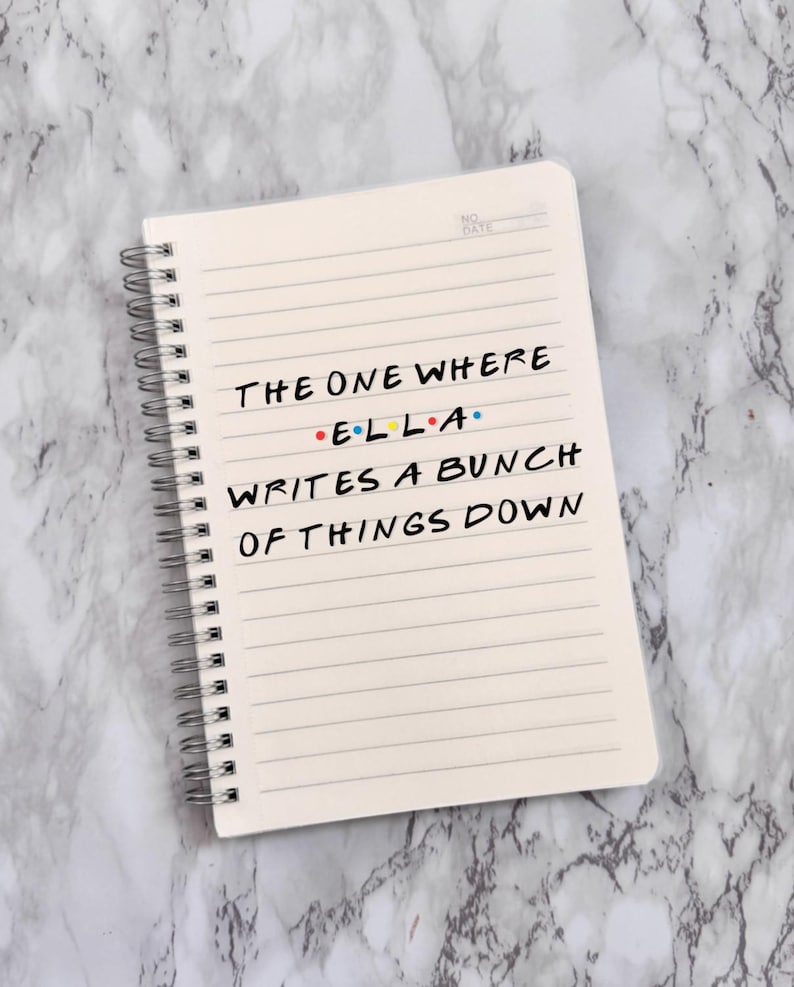 May include: A cream-coloured, spiral-bound notebook with lined pages. The text on the page reads "THE ONE WHERE E.L.L.A. WRITES A BUNCH OF THINGS DOWN" in black ink. The notebook is on a marble-effect surface.