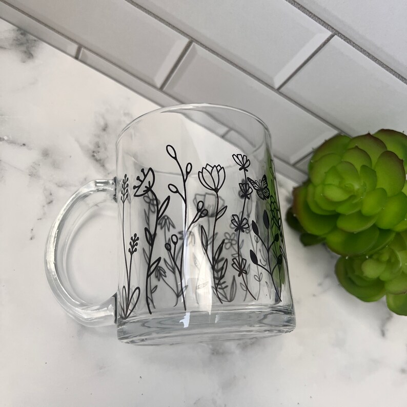 May include: A clear glass mug with a black floral design and a butterfly. The mug has a handle and is sitting on a white surface.