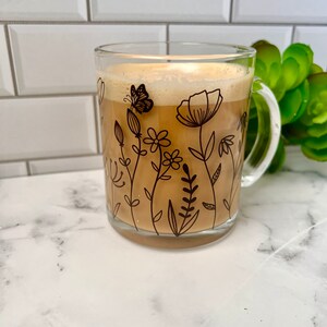 May include: A clear glass mug with a black floral design and a brown liquid inside. The mug has a handle and is sitting on a white marble surface.