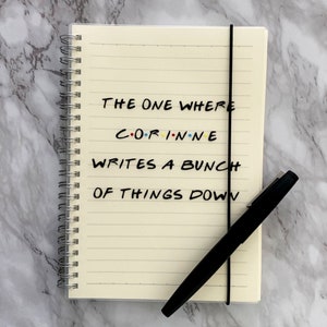 May include: Cream-coloured, spiral-bound notebook with lined pages and a black elastic band. The cover reads "THE ONE WHERE CORINNE WRITES A BUNCH OF THINGS DOWN" in black and colourful lettering. A black pen lies across the notebook.