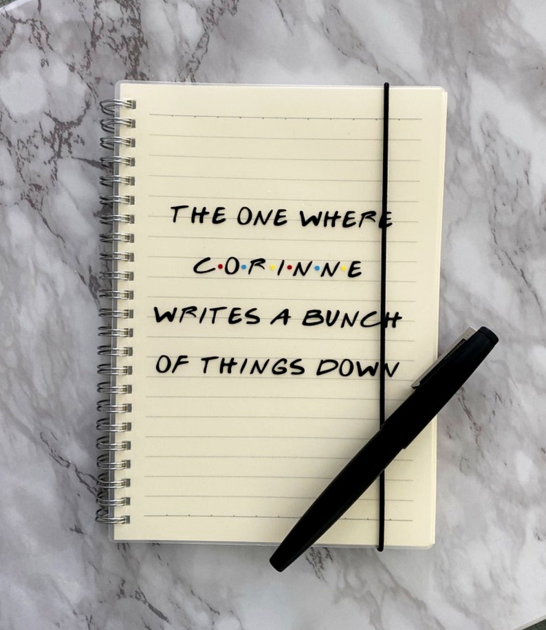 May include: Cream-coloured, spiral-bound notebook with lined pages and a black elastic band. The cover reads "THE ONE WHERE CORINNE WRITES A BUNCH OF THINGS DOWN" in black and colourful lettering. A black pen lies across the notebook.