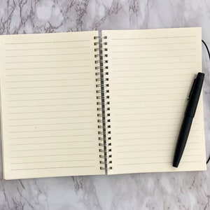 May include: An open notebook with lined cream-coloured pages, a silver spiral binding, and a black pen. The pen rests on the right page. The notebook is on a marble-effect surface.