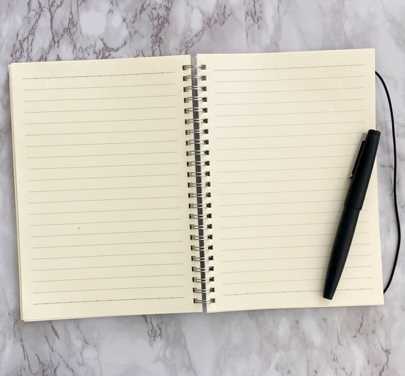 May include: An open notebook with lined cream-coloured pages, a silver spiral binding, and a black pen. The pen rests on the right page. The notebook is on a marble-effect surface.