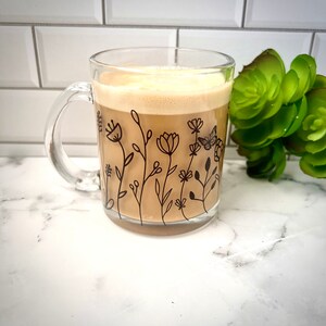 May include: A clear glass mug with a black floral design and a butterfly. The mug is filled with a light brown beverage.