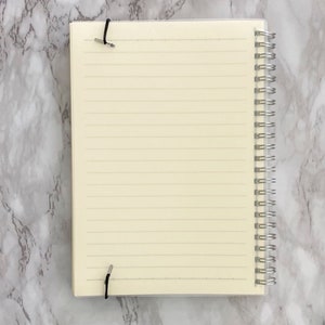 May include: A spiral-bound notebook with lined cream-coloured pages. The notebook is held closed with two black elastic bands. The spiral binding is silver. The notebook is open, revealing the lined pages.
