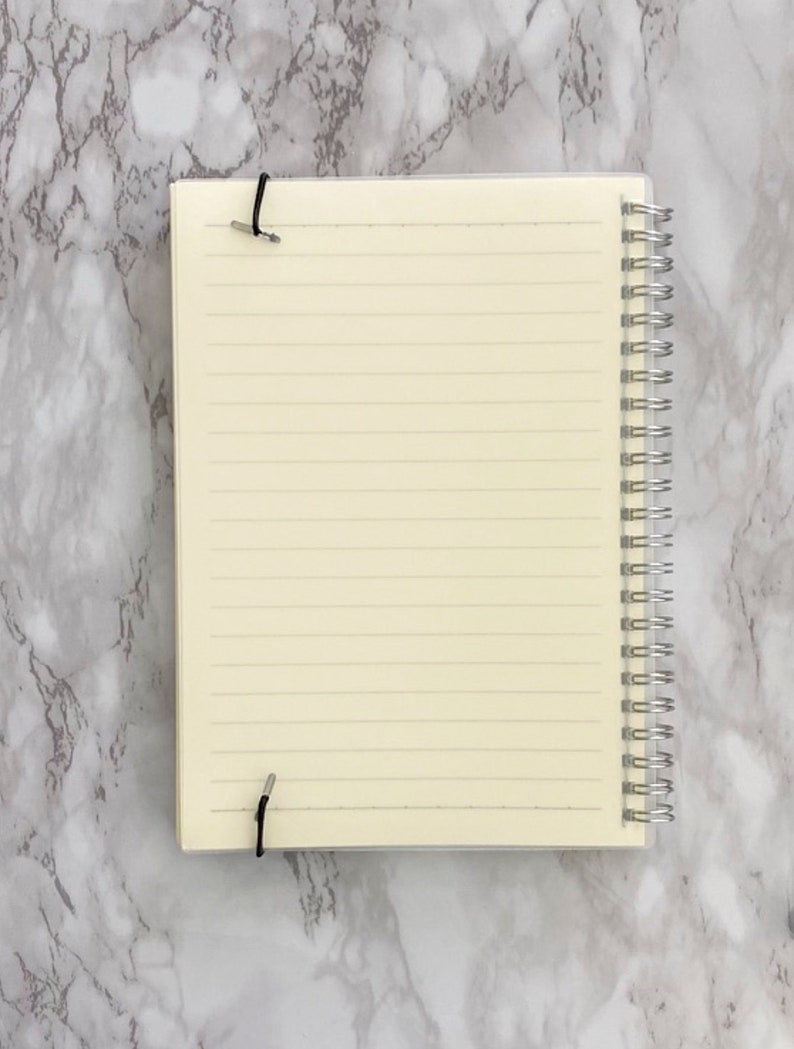 May include: A spiral-bound notebook with lined cream-coloured pages. The notebook is held closed with two black elastic bands. The spiral binding is silver. The notebook is open, revealing the lined pages.