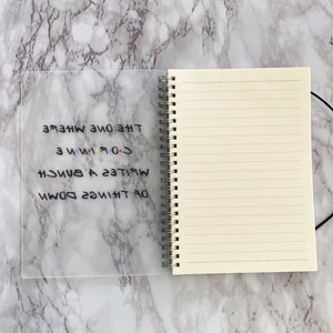 May include: A lined notebook with a spiral binding and a clear plastic cover. The cover displays the text "The One Where I Write A Bunch Of Things Down" in black, with the "Friends" logo.