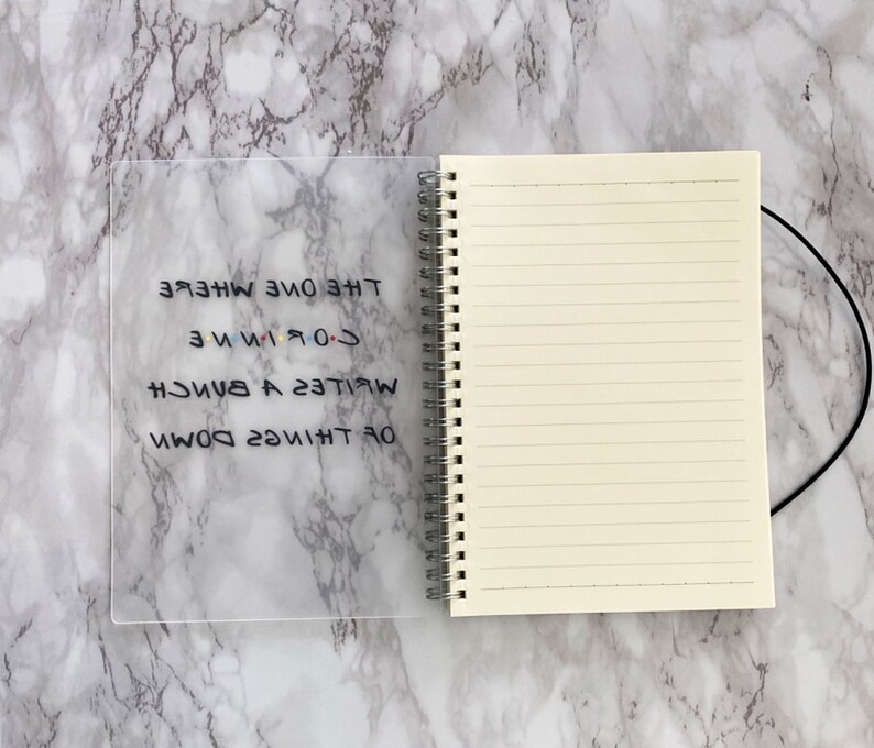 May include: A lined notebook with a spiral binding and a clear plastic cover. The cover displays the text "The One Where I Write A Bunch Of Things Down" in black, with the "Friends" logo.