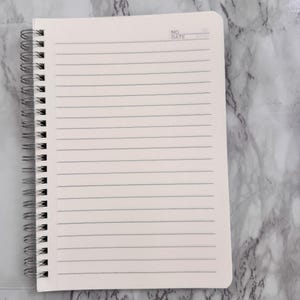 May include: A cream-coloured spiral-bound notebook with lined pages. The top right corner has the text "NO DATE". The notebook is open, revealing the ruled pages, and the spiral binding is silver.