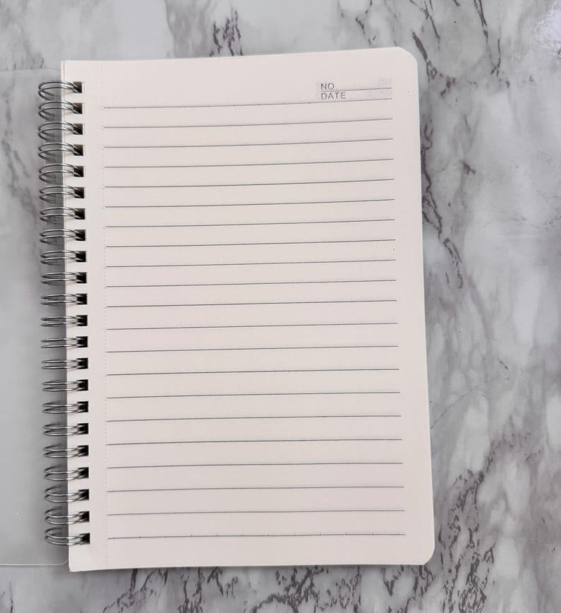 May include: A cream-coloured spiral-bound notebook with lined pages. The top right corner has the text "NO DATE". The notebook is open, revealing the ruled pages, and the spiral binding is silver.
