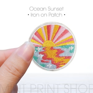 Könnte beinhalten: Runder Aufbügler mit einem Ozean-Sonnenuntergang-Design. Der Aufnäher hat einen weißen Rand und zeigt einen farbenfrohen Sonnenuntergang mit gelben, rosa und orangefarbenen Strahlen über einem blauen und roten Ozean. Der Text auf dem Aufnäher lautet "Ocean Sunset - Iron on Patch."
