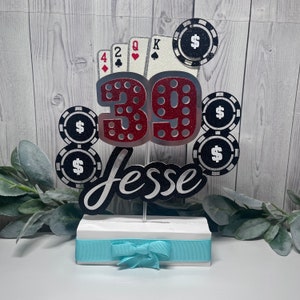 May include: A red and silver glitter cake topper with the number 39, the name Jesse, and playing cards and poker chips. The topper is on a white base with a light blue ribbon.