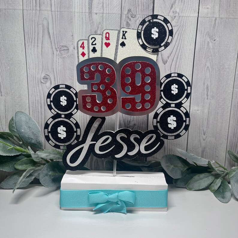May include: A red and silver glitter cake topper with the number 39, the name Jesse, and playing cards and poker chips. The topper is on a white base with a light blue ribbon.