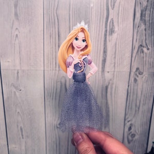 May include: A princess-themed cake topper featuring a cartoon illustration of a princess with long blonde hair, a silver crown, and a purple and gray dress. The princess is holding her hand on her hip. The dress has a tulle skirt.